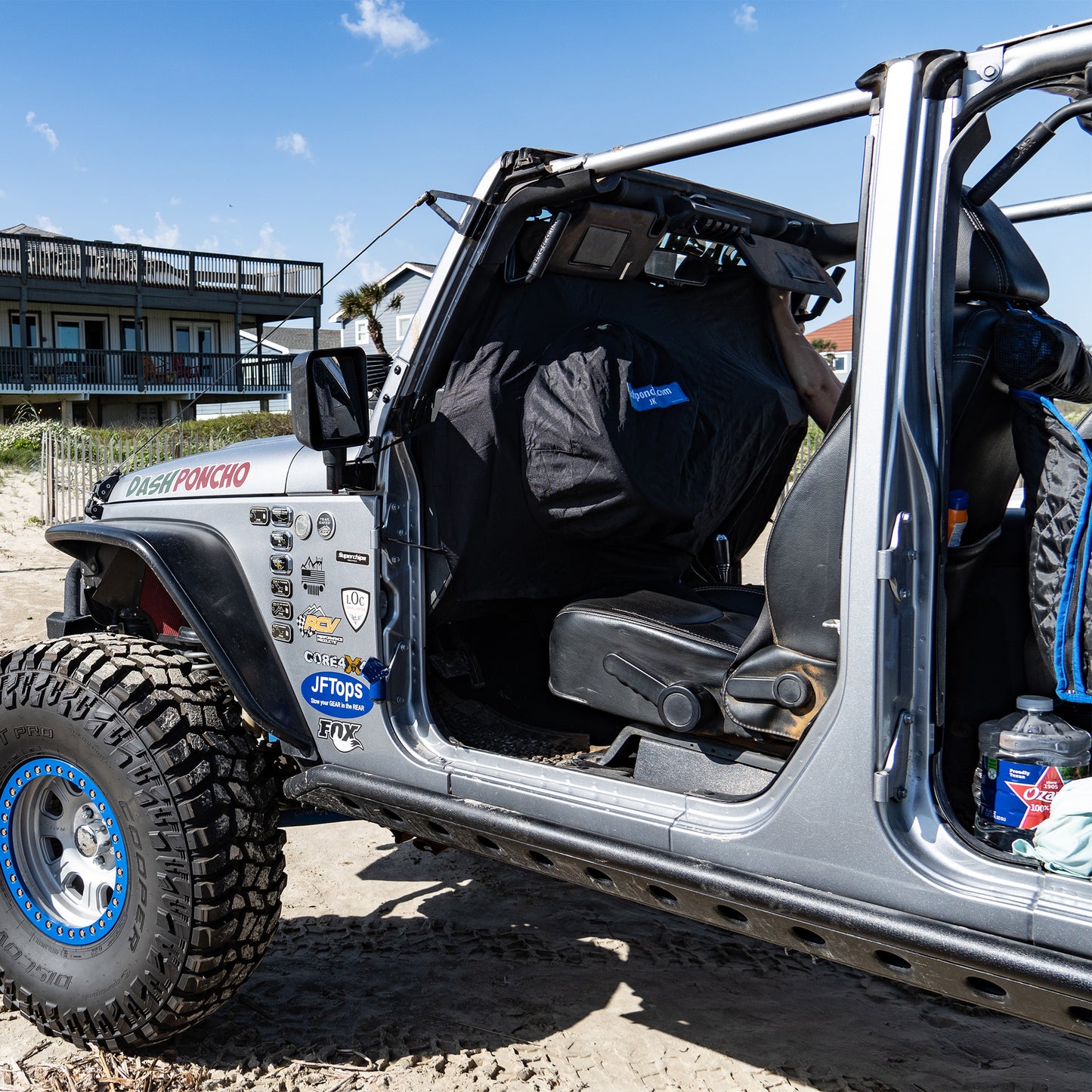 Open door of a silver off-road vehicle with JKU JEEP WRANGLER RAIN COVER on the Beach with  clear sky and building in the background